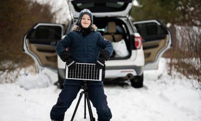 Boy with solar panel battery on tripod against car in winter woods.