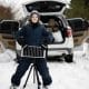 Boy with solar panel battery on tripod against car in winter woods.